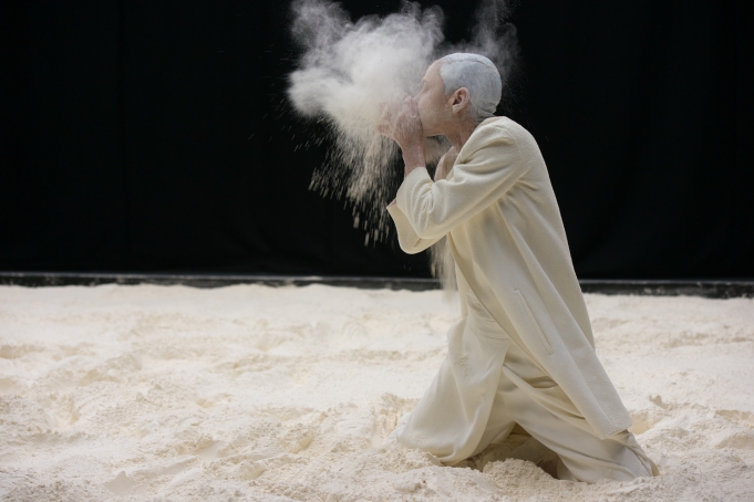 Image of actor in white on knees blowing white dust. Photo: Warren Orchard