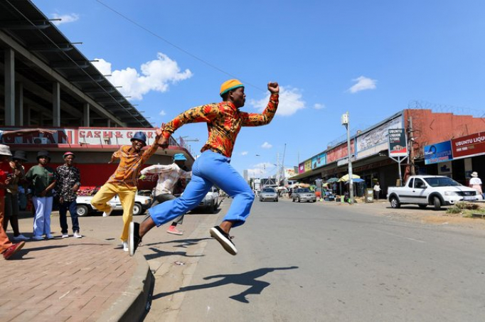 A black man does a running jump in a city street with a bright blue sky