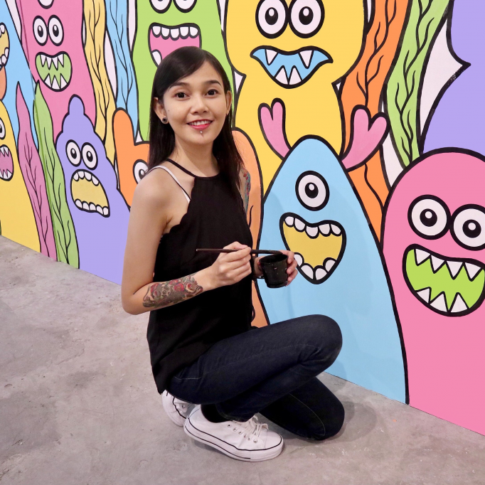 A woman kneels in front of a bright wall painting, holding a brush and smiling